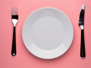 Blank flat plate, knife, fork on a color pink table, top view.