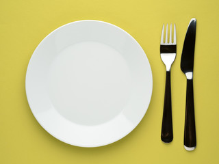 Blank flat plate, knife, fork on a color yellow table, top view.