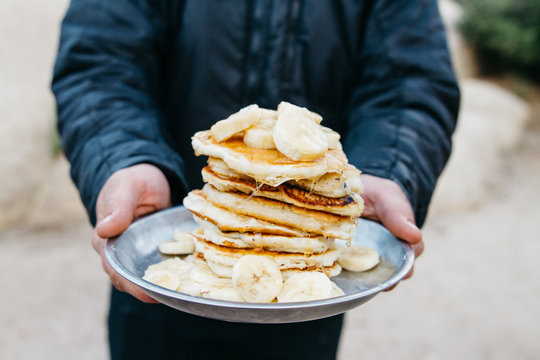young man holds pile of pancakes on a plate while camping