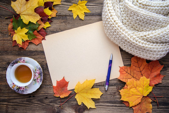 Autumn picture of yellow leaves, a cup of tea, a scarf and a piece of paper with pen on wooden background