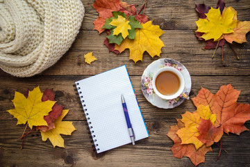 Autumn picture of yellow leaves, a cup of tea, a scarf and a piece of paper with pen on wooden background