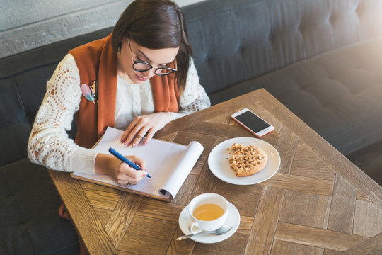Young businesswoman in glasses and white sweater is sitting in cafe at table, signing documents. Student is studying online, doing homework. On table cup of tea, cookies, smartphone. E-learning.