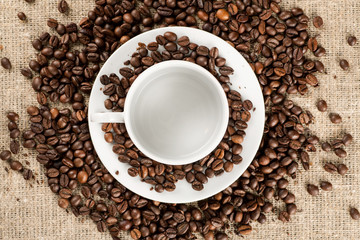 White coffee mug with plate among many of roasted coffee beans, top view