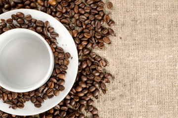 Fototapeta na wymiar White coffee mug with plate among many of roasted coffee beans, top view, text space