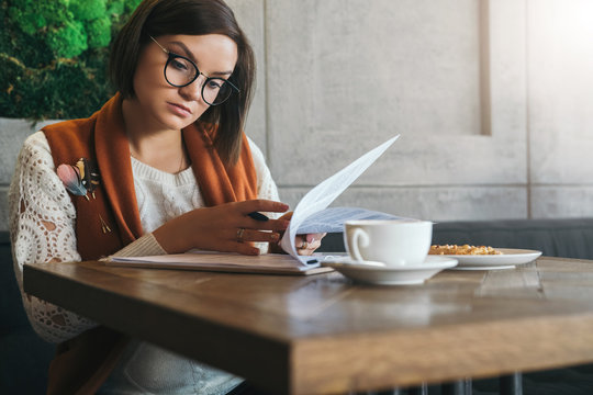Front view.Young businesswoman is sitting in coffee shop at table,reading documents.Student is studying online, doing homework.On table cup of tea, cookies. E-learning, education. Freelancer working.