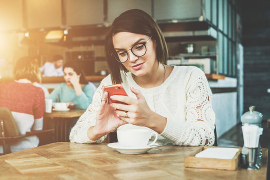 Young businesswoman in glasses and white sweater is sitting in cafe at table and using smartphone, working. Student is studying online, e-learning. Online marketing, education. Hipster girl chatting.