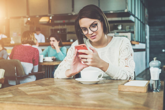 Young happy businesswoman in glasses and sweater is sitting in cafe at table and using smartphone, working.E-learning,online marketing,education.Hipster girl looking on screen of phone,checking email.