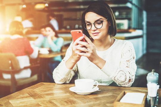 Young businesswoman in glasses and white sweater is sitting in cafe at table and using smartphone, working.E-learning,online marketing,education.Hipster girl looking on screen of phone,checking email.