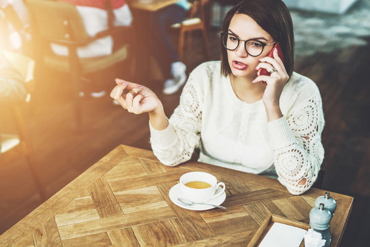 Young businesswoman in glasses and white sweater is sitting in cafe at wooden table and talking on mobile phone. Telephone conversations. Hipster girl relaxing, drinking tea, calling her friends.