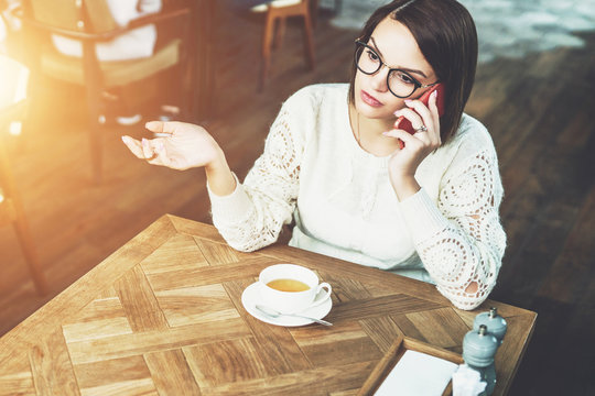 Young businesswoman in glasses and white sweater is sitting in cafe at wooden table and talking on cell phone. Telephone conversations. Hipster girl freelancer discusses working questions over phone.