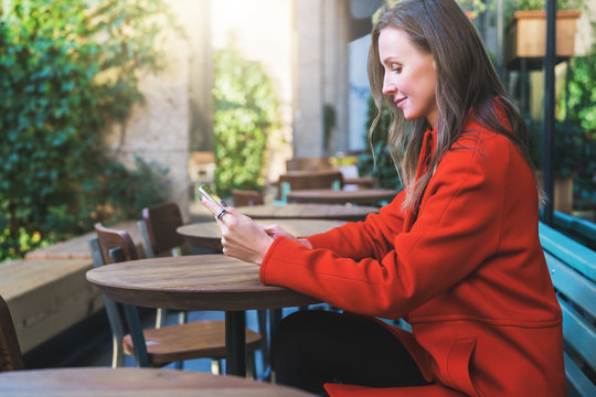 Side view.Young smiling attractive woman in orange coat is sitting outside in cafe at table and uses tablet computer. Girl checking email, blogging, chatting, reading e-book.Social media, e-learning.