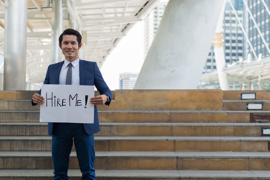 Hire me! Handsome businessman wear blue suit holding poster with hire me text message while standing outdoors and against building structure