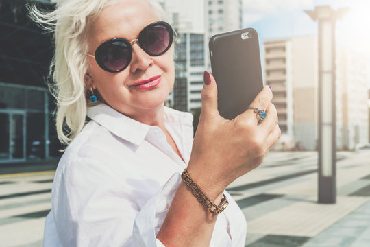 Summer day. Middle-aged woman, a pensioner, dressed in a white shirt and sunglasses, stands on a city street and uses a smartphone. In the background is a modern glass building. Social media, network.