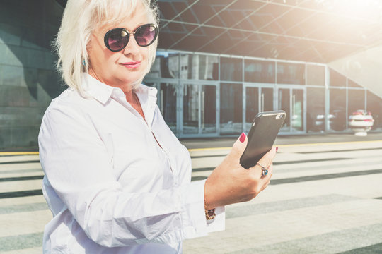 Summer sunny day. A middle-aged woman, a pensioner, dressed in a white shirt and sunglasses, stands on a city street and uses a smartphone. In the background is a modern glass building. Lady chatting.