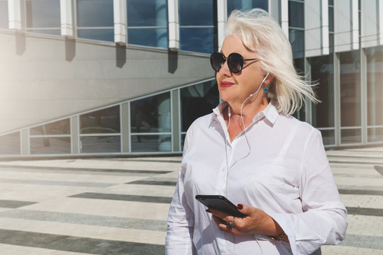 Summer sunny day.Middle-aged woman,retired woman dressed in white shirt and sunglasses,stands on city street,holds smartphone and listening music in headphones. In background is modern glass building.