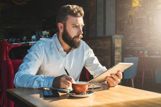 Young bearded businessman is sitting in cafe at table, holding tablet computer and writing in notebook. On desk is cup of tea and smartphone.Online education, e-marketing, e-learning. Film effect.