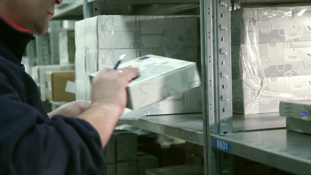 Man Storekeeper takes goods from shelf warehouse of auto parts and makes a mark on paper. waybill in the hands of the worker draws up a packed box for delivery to buyer.