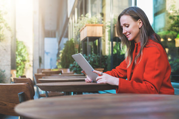 Side view.Young smiling attractive woman in orange coat is sitting outside in cafe at table and uses tablet computer. Girl checking email, blogging, chatting, reading e-book.Social media, e-learning.