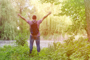 Fototapeta na wymiar Guy with a backpack is standing on the shore of a forest lake with arms outstretched, a back view