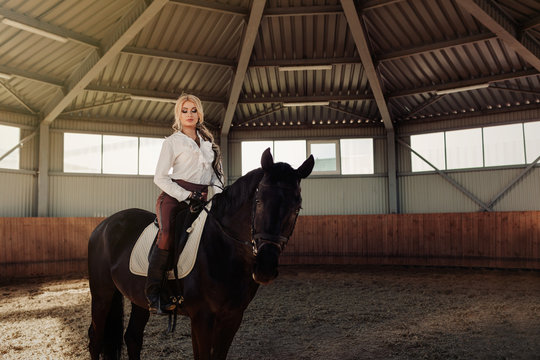 Beautiful elegant young blonde girl sits on a her black horse dressing uniform competition white blouse shirt and brown pants.