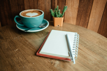 cup with coffee has a heart shape on top and diary notebook with pen on top placed on wooden table . wood wall are background. image for business, coffee art, beverage, drink, food, art concept