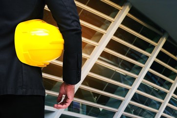 back view of engineer and holding yellow safety helmet standing with exterior of glass modern office building at business center background, industrial and business concept