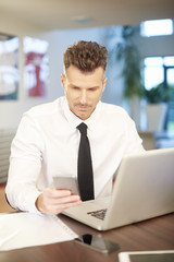 Businessman working at the office. Shot of a middle aged businessman using his mobile phone and laptop while working at the office. 