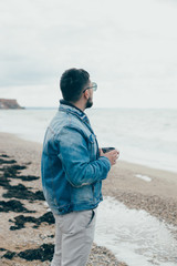 Stylish young man with a beard standing near ocean with a cup of tea