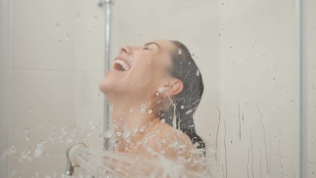 Laughing woman has fun in the shower cabin and pours the water on the glass