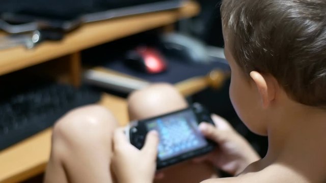 Child is playing in a portable game console sitting on a chair at home. Focus is on the boy. 8 years old kid playing video games. Child using console.