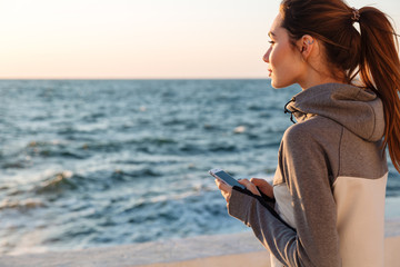 Naklejka premium Back view of pretty sport woman holding smartphone, looking at the sea