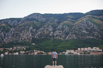 Young tanned man in shorts and a T-shirt is standing on the pier and looking at the sea with mountains in the background. 