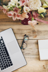 Office workplace essentials: laptop, paper notebook, glasses and a bouquet of flowers on the rustic wooden table background