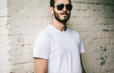 Obraz premium Young stylish man wearing white blank t-shirt with beard in glasses, standing on the street on city background. Street photo