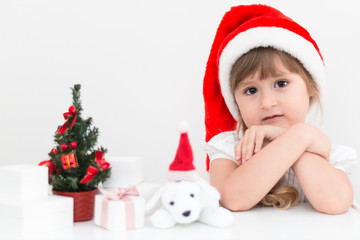 Little cute girl wearing a Santa Claus hat is sitting next to a little Christmas tree with a bunch of gifts and a plush dog. Christmas and New Year card. 2018 year.