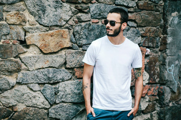 Young stylish man wearing white blank t-shirt with beard in glasses, standing on the street on city...