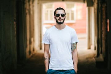 Young stylish man wearing white blank t-shirt with beard in glasses, standing on the street on city background. Street photo