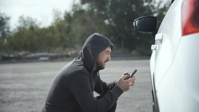 Thief wearing hoodie hacking car using smartphone to open door and steal suitcase