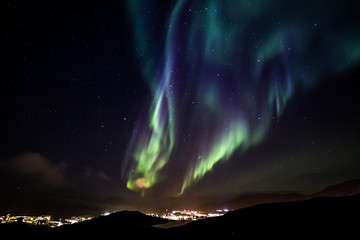 Glows of Northern Lights with shining stars on the sky over the mountains and highlighted city, Nuuk, Greenland