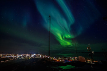Green waves of Aurora Borealis with shining stars over the fjord and highlighted city, Nuuk, Greenland