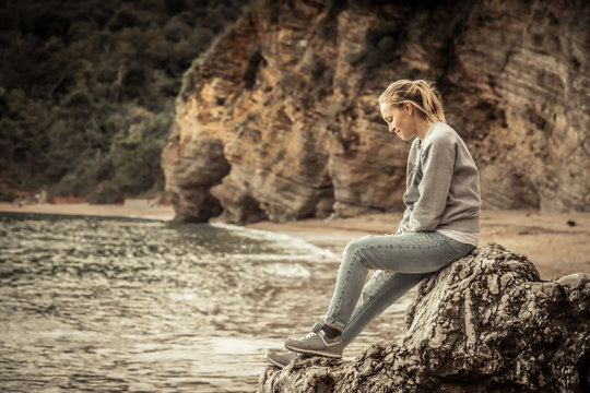Pensive lonely young woman traveler relaxing on a big cliff stone on the beach looking at wild mountain scenery in retro vintage style