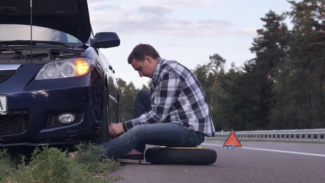 Caucasian man changing a tire sitting on stepney near car on the roadside. Handsome traveller outdoors