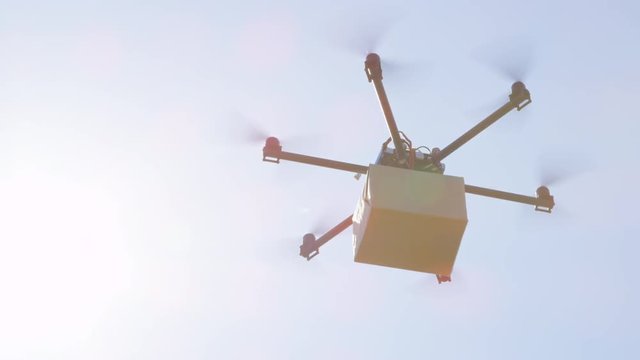 CLOSE UP LENS FLARE: UAV drone delivery. Multicopter flying brown package over sun. Drone delivering goods to your home. Futuristic shipment by helicopter drone. Multirotor logistics and transport