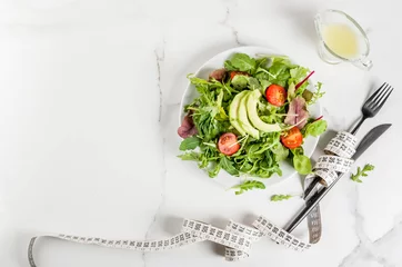 Foto op Plexiglas Healthy balanced diet concept, weight loss, calorie counting. Plate with green salad leaves, tomatoes, avocado with yogurt dressing, white table, with fork, knife, measuring tape, top view copy space © ricka_kinamoto