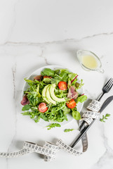 Healthy balanced diet concept, weight loss, calorie counting. Plate with green salad leaves, tomatoes, avocado with yogurt dressing, white table, with fork, knife, measuring tape, top view copy space