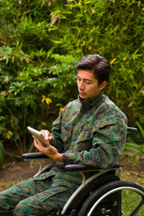 Close up of a handsome soldier sitting on wheel chair using his tablet with both hands, and wearing military uniform in a nature background