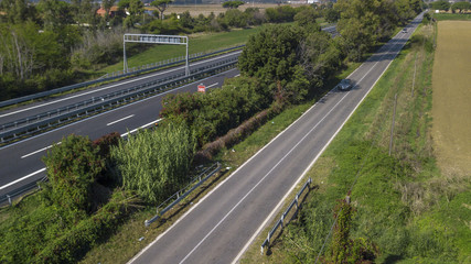 Vista aerea di un tratto di autostrada italiana. Ci sono due corsie in ogni carreggiata per entrambi i sensi di marcia. Al suo lato una strada piccola e secondaria.