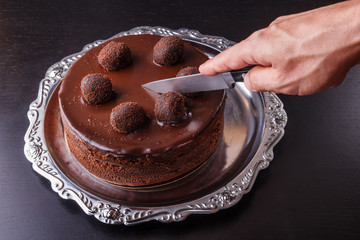 A man's hand with a kitchen knife above a chocolate homemade biscuit cake on a metal plate