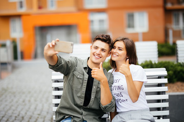 Handsome young couple sitting on the bench in the center of the city and taking a selfie with thumbs up. Love and tenderness. Lifestyle concept