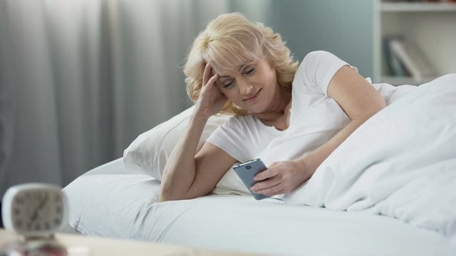 Smiling mature woman lying in bed and checking e-mail on smartphone, morning